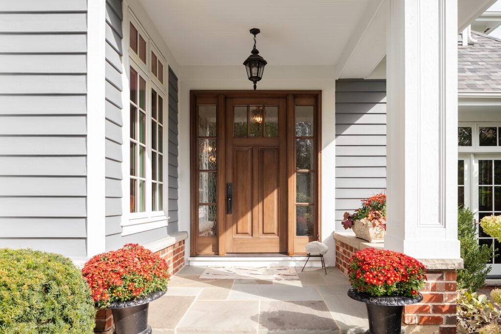 A front door and covered porch detail on a home with grey siding, white trim, red brick, a wooden front door.