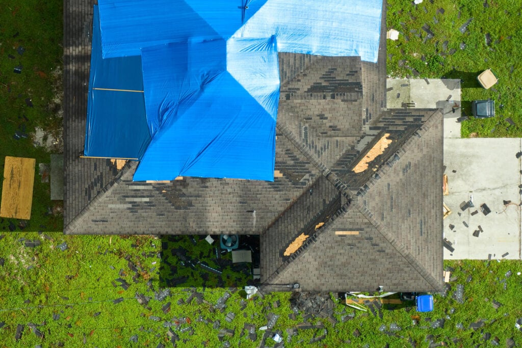 hail damage roof repair-aerial view of storm damaged roof with debris scattered in yard and roof partially covered in blue tarp