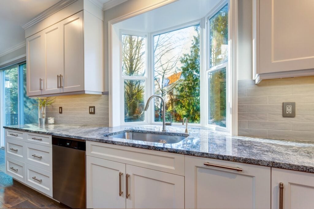Luxury home interior boasts amazing white kitchen with custom white shaker cabinets topped with granite fitted with a wide stainless steel sink and pull out faucet placed under the window.
